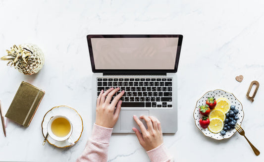 Eine Frau tippt auf einem Laptop mit weißen Bildschirm. Links steht eine Teetasse mit einem gelben Tee, eine Dekoananas und ein Notizbuch mit Stift. Rechts davon ein Obstteller.