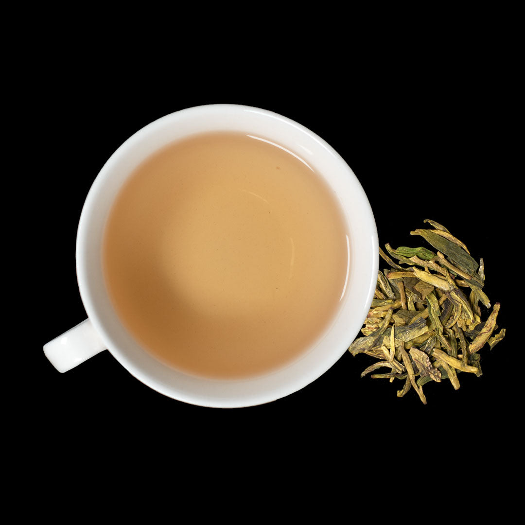 Eine weisse Tasse voller Tee in gelber Farbnote mit Henkel und rechts davon loser Drachenbrunnentee.