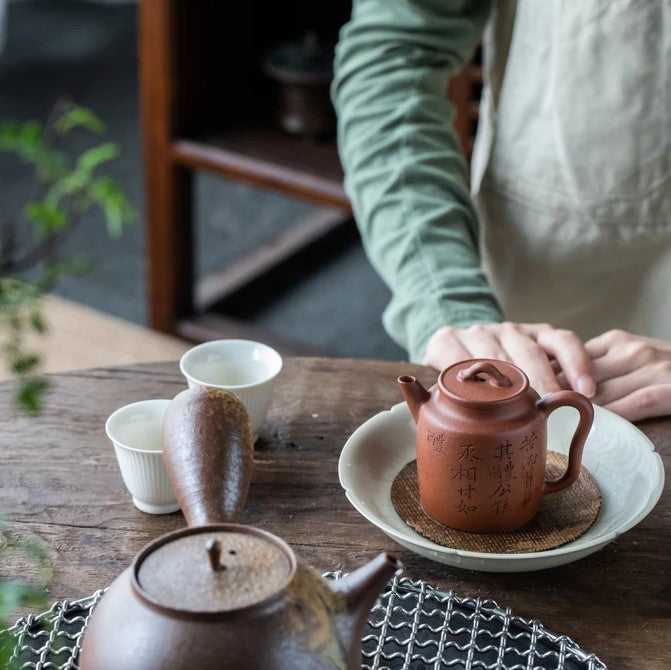 Das Bild zeigt eine Teezubereitung, die von einer Frau mit gepflegten Fingernägeln zelebriert wird. Es sind 2 Kannen zu sehen, womit die Frau den Tee zubereitet und in einer Tasse einschenken kann.