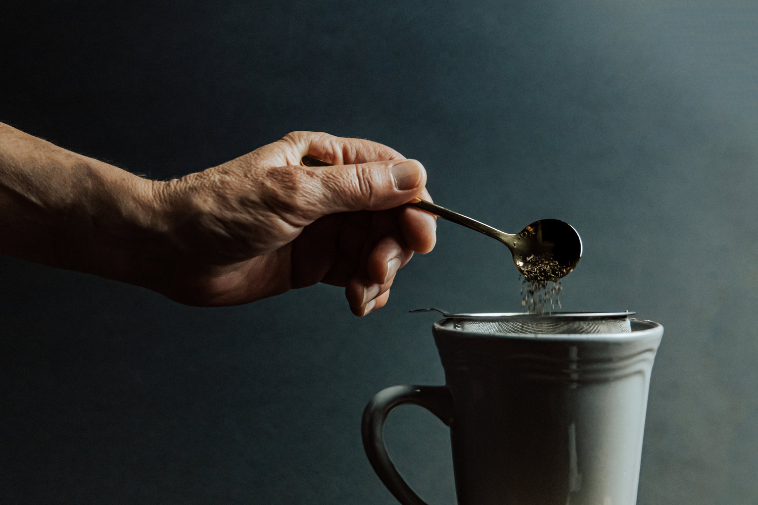 Eine Person füllt mit einem Teelöffel den Grüntee in ein Teesieb aus Metall, welches sich in einer grünen Tasse befindet