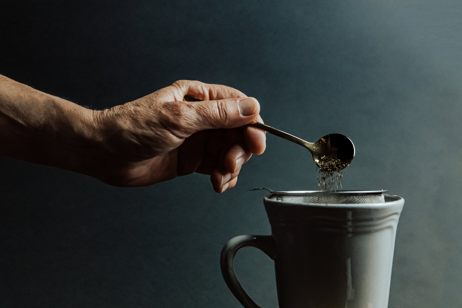 Eine Person füllt mit einem Teelöffel den Grüntee  in ein Teesieb aus Metall, welches sich in einer grünen Tasse befindet