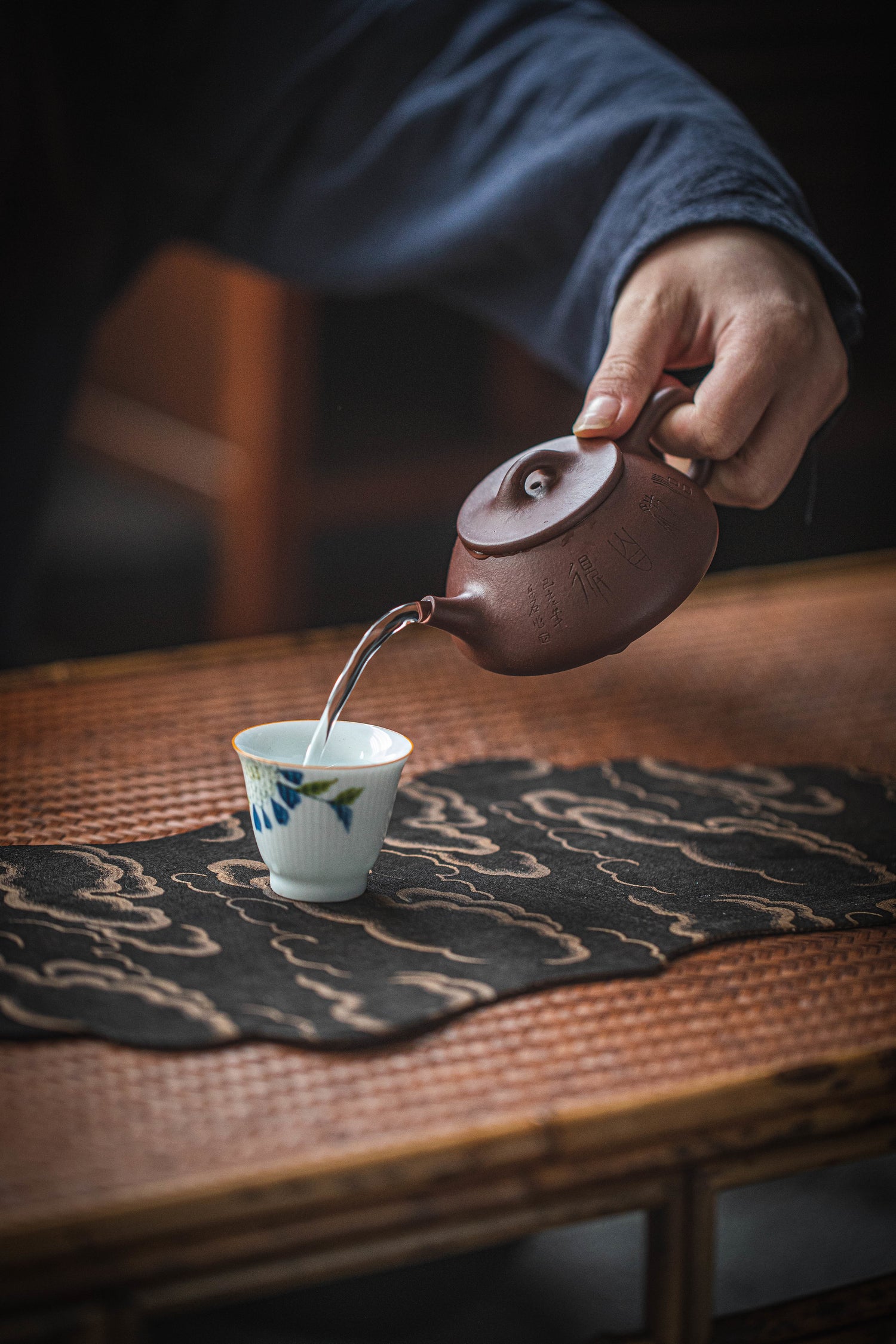 Ein asiatisch wirkendes Bild, wo eine Person Tee aus einer Gußeisenkanne in einen kleinen Teebecher eingiesst. Der Teebecher steht auf einem Basttisch mit einer schönen Decke mit schwarzem Hintergrund und beige/goldenen Wolken